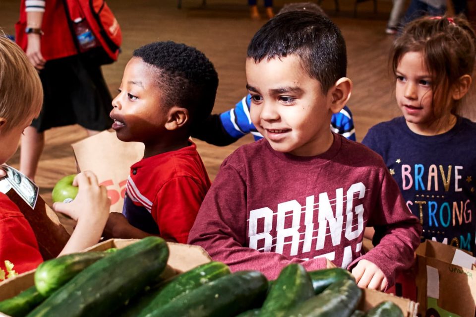 Young children stand in front of a table filled with fresh produce during a school activity