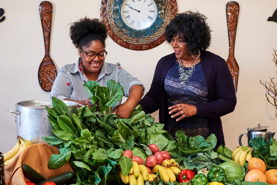 Two Black women arrange a cornucopia of fresh produce on a table