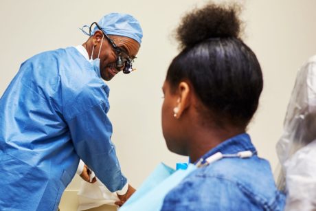 A Black dental professional wearing scrubs converses with a Black girl patient in an examination room.
