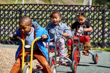 Three young Black children ride tricycles in a playground blacktop