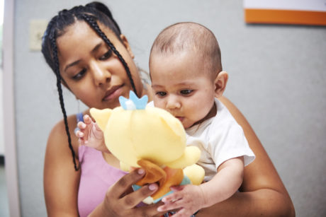 A mother with two small braids holds an infant, both who are people of color, while they play with a yellow plush toy