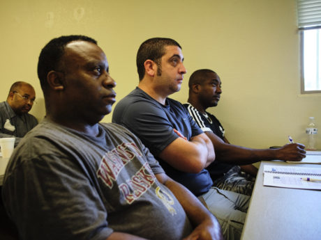 A row of men with different ages and racial backgrounds watch a presentation and take notes during a focus group session