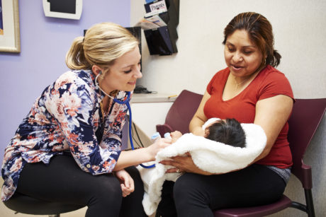 A mother, who is a person of color, holds her infant in a fluffy white blanket while a white pediatrician uses a stethoscope