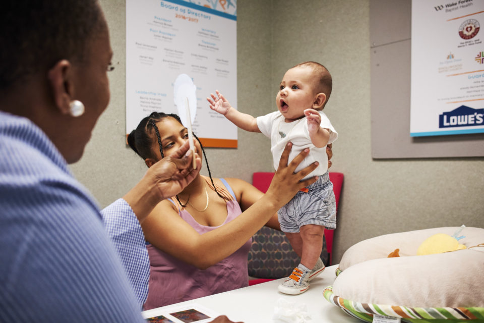 A mother, who is a person of color, holds her infant upright while a Black woman doctor tests their facial recognition ability