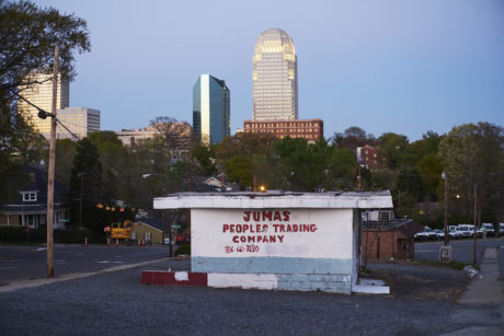 A small abandoned building with signage for the "Jumas Peoples Trading Company" stands in the foreground of a cityscape of Winston-Salem at dusk