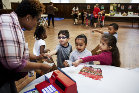 A Black woman teacher leads a group of young Black and Brown students through a mock banking activity in a gymnasium