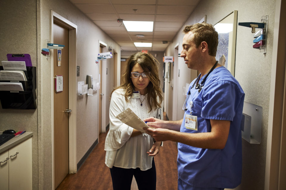Two white clinicians, one wearing a white top and the other blue scrubs, talk in the hallway of a medical office