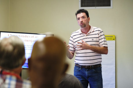 A white man wearing a striped shirt gives a slide presentation in front of a focus group