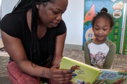 A Black woman with long braids reads a picture book with a young Black child in a colorful early learning classroom.