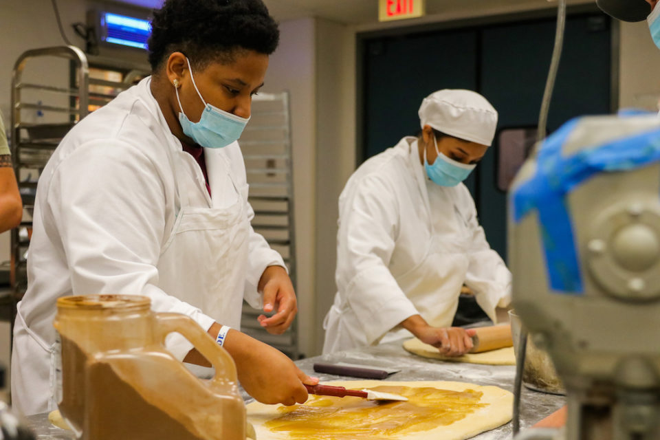 Two youth wearing chef uniforms and face masks prepare dough in a teaching kitchen