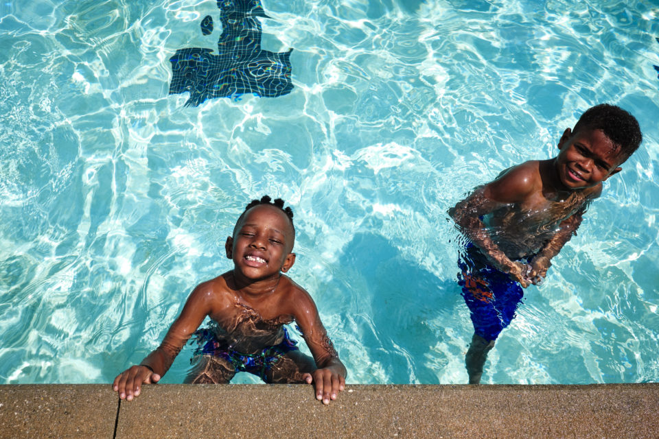 Two young Black boys grin at the viewer while swimming in the shallow end of a pool in a sunny day