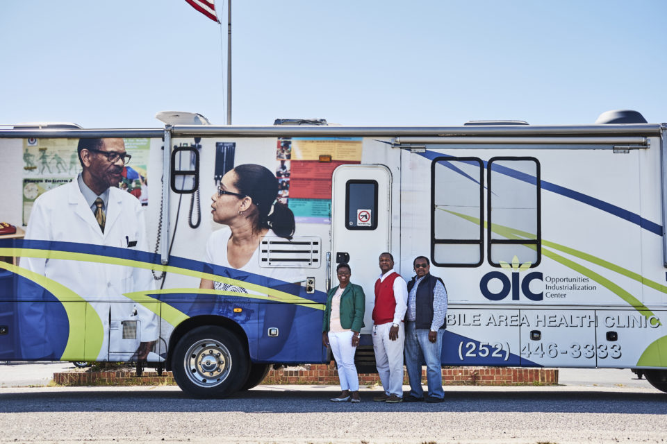 Three Black adults wearing casual office attire and bold colored vests stand in front of the OIC mobile clinic